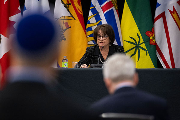 Deborah Lyons s'exprimant à un pupitre avec des drapeaux canadiens en arrière-plan et en avant-plan, les têtes de deux membres du public, dont l'un porte une kippa