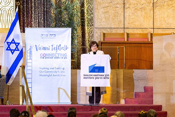 Deborah Lyons s’exprime à un pupitre affichant « Shalom Hartman Institute », à côté d'un drapeau israélien et d'une affiche sur laquelle est écrit « Va'ani Tefilali : Connecting The Divine Within Us »