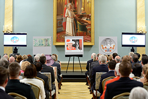 Seated people look at items displayed on easels placed on a platform. A portrait of Queen Elizabeth II hangs on a wall in the background.