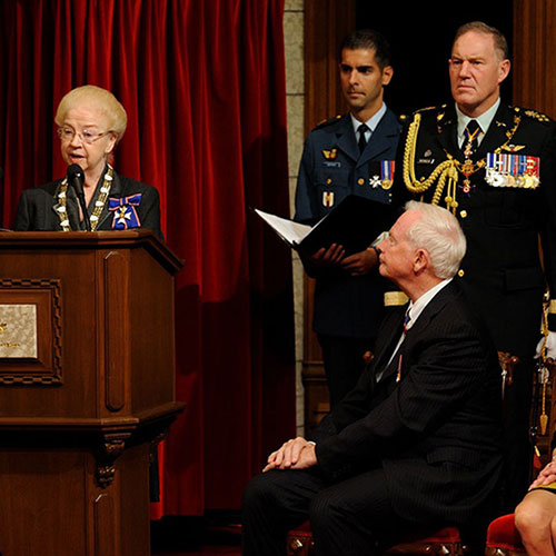 A woman speaking at a podium. David Johnston and Sharon Johnston are seated and listening.