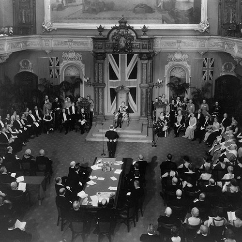 William Lyon Mackenzie King facing away from the camera and speaks to Lord Tweedsmuir and others in the Senate Chamber.