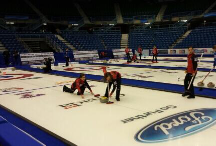 Des personnes jouent au curling. Sur la glace apparaît le mot-symbole Canada accompagné des logos et identificateurs d'autres bailleurs de fonds.