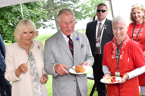 The Prince of Wales and the Duchess of Cornwall standing next to a woman in a red shirt, holding a plate with a dessert.