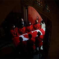 At the centre of this dark, shadowed image, we see the casket covered by the National Flag of Canada being carried  by RCMP pallbearers in red serge through an arched doorway. 