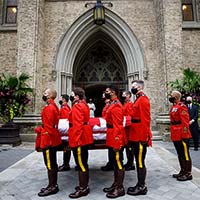 Eight RCMP pallbearers in red serge are seen holding the casket while another one directs them. A priest is seen standing in the doorway of the cathedral.