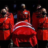 A front view of the casket covered with the National Flag of Canada being carried by RCMP pallbearers in red serge. They are wearing black masks.