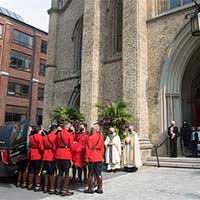 RCMP in red serge exit the cathedral carrying a casket, which is covered with the National Flag of Canada. The clergy and the family are seen standing at the entrance of the cathedral.