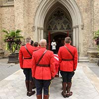 RCMP pallbearers in red serge are seen carrying the casket, which is covered with the National Flag of Canada, in the cathedral courtyard. A priest is seen standing in the doorway of the church.