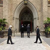 Six hommes en costume portant un masque, marchent sous l’arche de la façade extérieure de la cathédrale. Les hommes maintiennent une distance physique entre eux. 