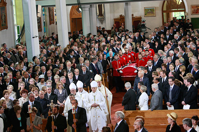 Led by the clergy, the military pallbearers carry the casket in a procession through the centre aisle of the church. Funeral guests stand in respect. 
