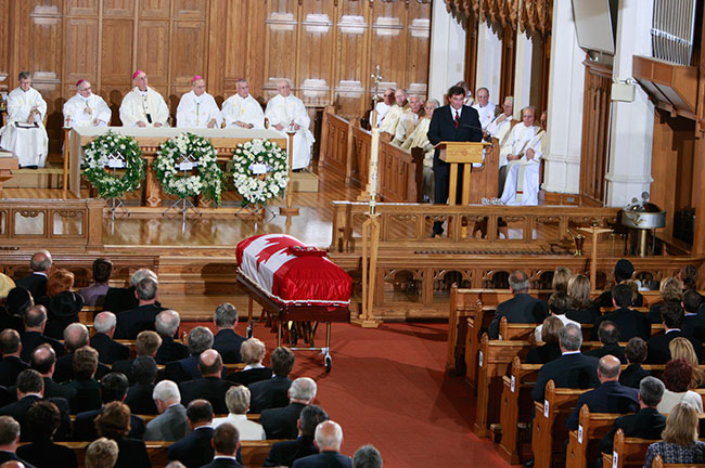 The casket draped with the National Flag of Canada and the insignia of the deceased rests at the front of the church. The adult son of the deceased delivers a eulogy from the lectern to the seated congregants and clergy.