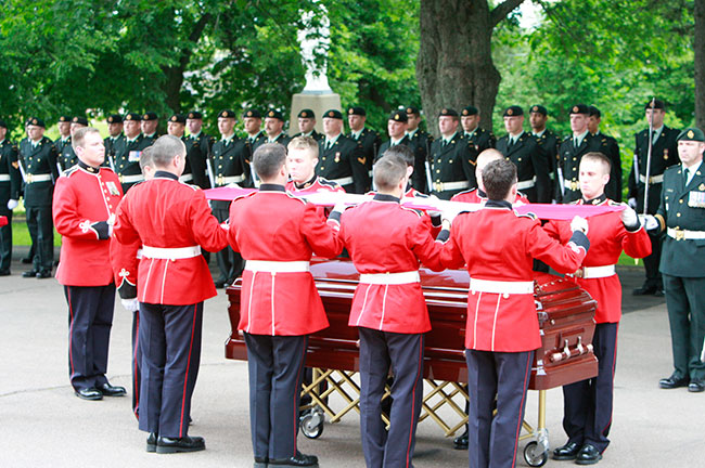 Les porteurs militaires du cercueil se préparent à plier le drapeau drapant le cercueil du défunt et devant être présenté à la famille. En arrière-plan, des membres de la garde d’honneur se tiennent au garde-à-vous.