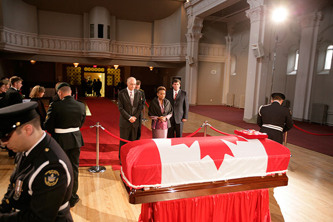Le cercueil, drapé du drapeau national du Canada et de l’insigne du défunt, repose à l’Institut de Memramcook. On aperçoit trois sentinelles à leurs postes veillant sur le défunt. Des personnes endeuillées lui rendent hommage.
