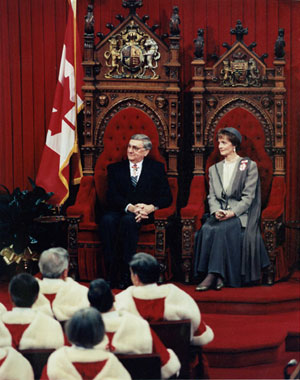 Governor General Roméo LeBlanc and his wife, Diana Fowler LeBlanc are seated on the throne seats in the Senate Chamber. Supreme Court Justices in red ceremonial robes are seated in the front centre aisle facing Their Excellencies.