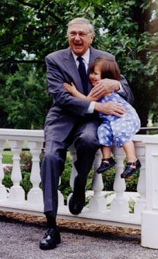An outdoor snapshot of Romeo LeBlanc in a suit and tie, hugging a female child in a blue dress, his goddaughter, Emma Kinsella.