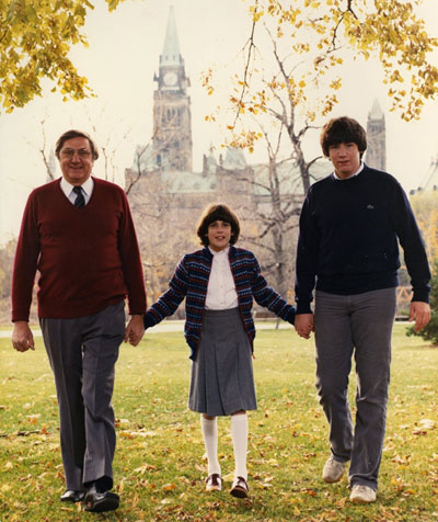 An outdoor autumn snapshot of Roméo LeBlanc, his young daughter and son holding hands walking forwards with the Parliament Buildings in the background.