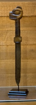Vertical display of a dagger with a steel blade, hide wrapping, and wooden pommel depicting the profile view of a bear’s head.