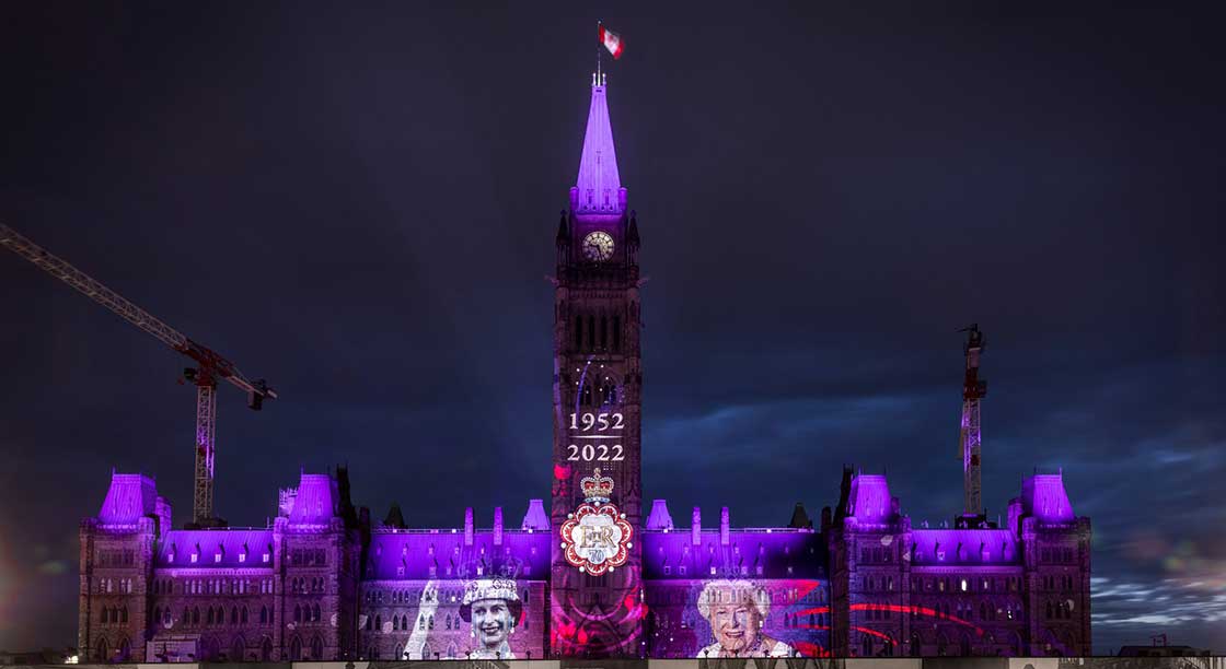 The Centre Block is lit up in purple. 2 photos of The Queen and one of the Canadian Platinum Jubilee Emblem are projected on the building.