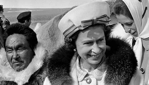 The Queen is smiling. She is wearing a hat and a fur trimmed parka.