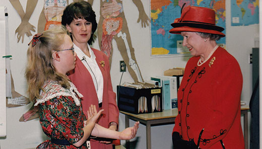 The Queen is standing in a classroom, smiling and talking with a young student as her teacher looks on.