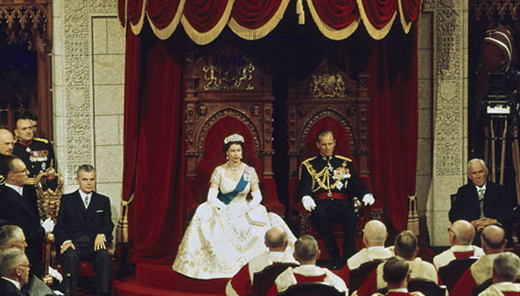 The Queen is sitting in the monarch's throne, in the Senate Chamber. The Duke of Edinburgh is to her left and John Diefenbaker to her right.