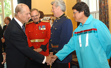 Prince Philip and a man woring an Indigenous traditional vest are shaking hands.