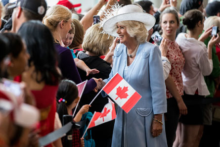 The Queen Consort is smiling as she shakes hands among a large crowd in front of her. Some people are waving small Canadian flags.