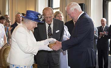 The Queen, wearing a blue hat and white dress receives the Sapphire Jubilee Snowflake Brooch from former Governor General David Johnston, while the Duke of Edinburgh looks on.
