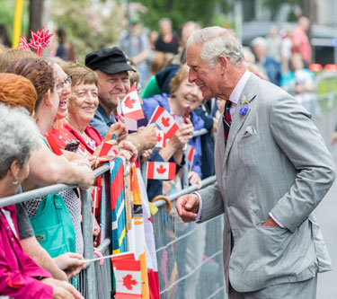 King Charles III is smiling to a crowd behind a fence. Members of the crowd hold a Canadian flag in their hands.