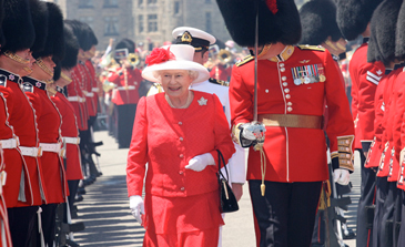 Queen Elizabeth II walks outside between 2 rows of ceremonial guards. A ceremonial guard also walks beside her.