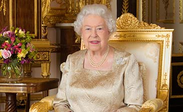 The Queen is wearing a golden dress with the Canadian diamond maple leaf brooch and triple string of pearls; she is sitting on a golden chair.