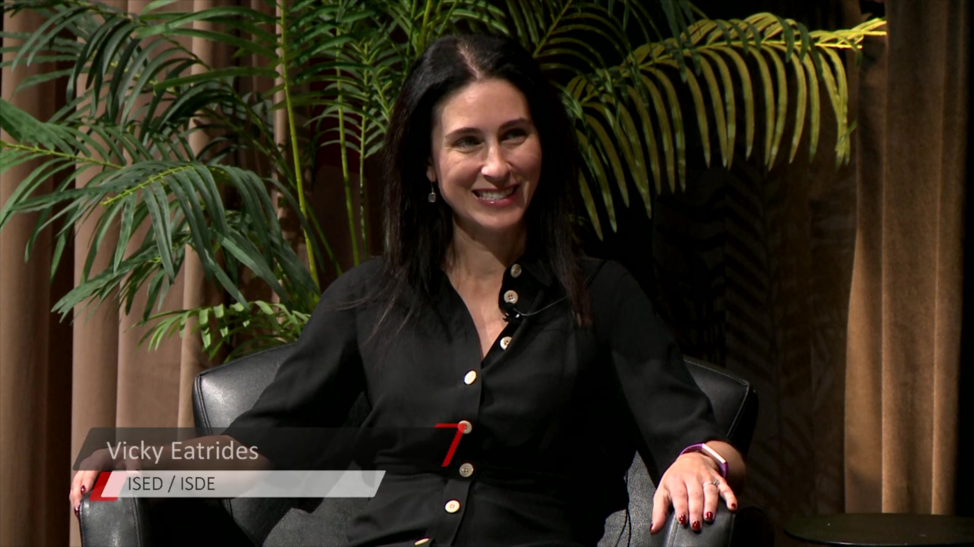 A woman, Vicky Eatrides, is seated in a black chair with green plants in the background