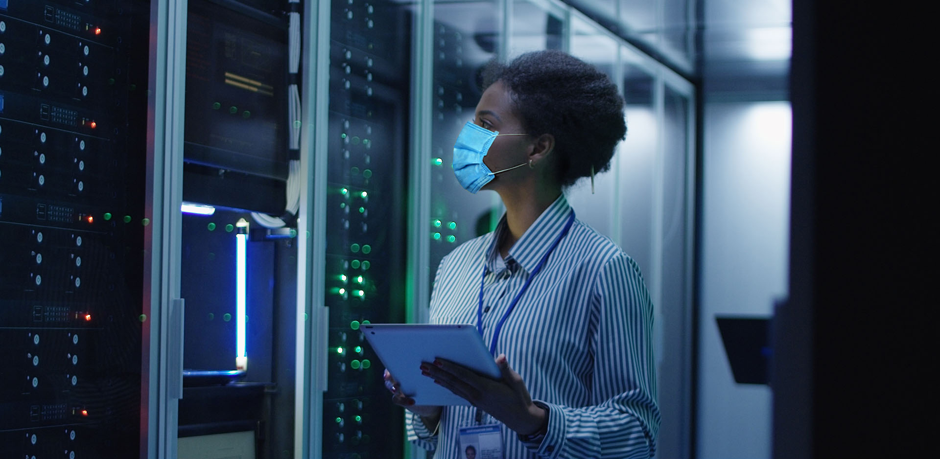 A masked individual, holding a clipboard, stands in front of a wall of computer servers