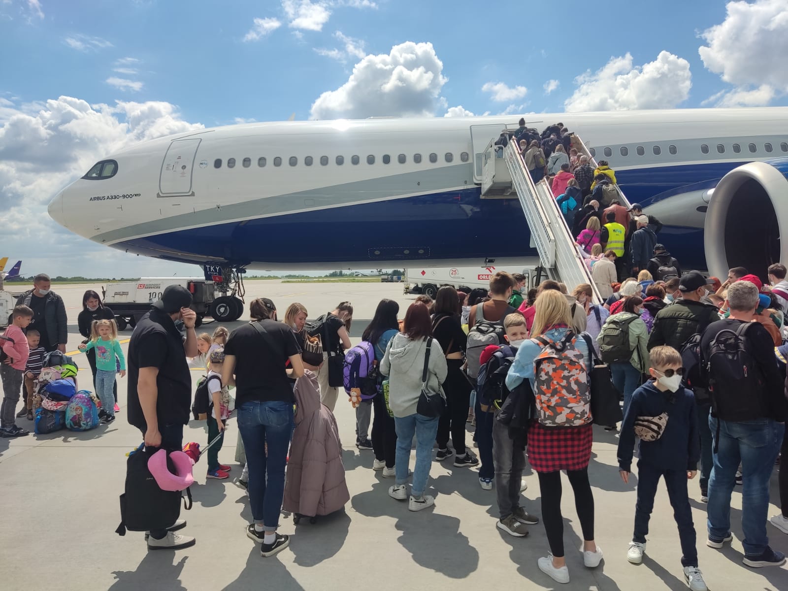 A crowd of people stands waiting with their luggage on a tarmac as others walk up a set of stairs and board a plane