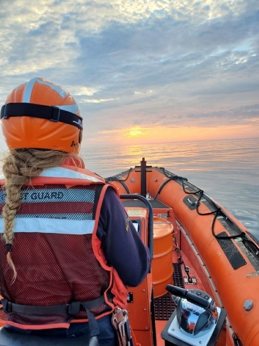 A person with their back to the camera wears a helmet and an orange vest that says “Coast Guard” on the back. They are steering an orange boat on the water towards the sunset