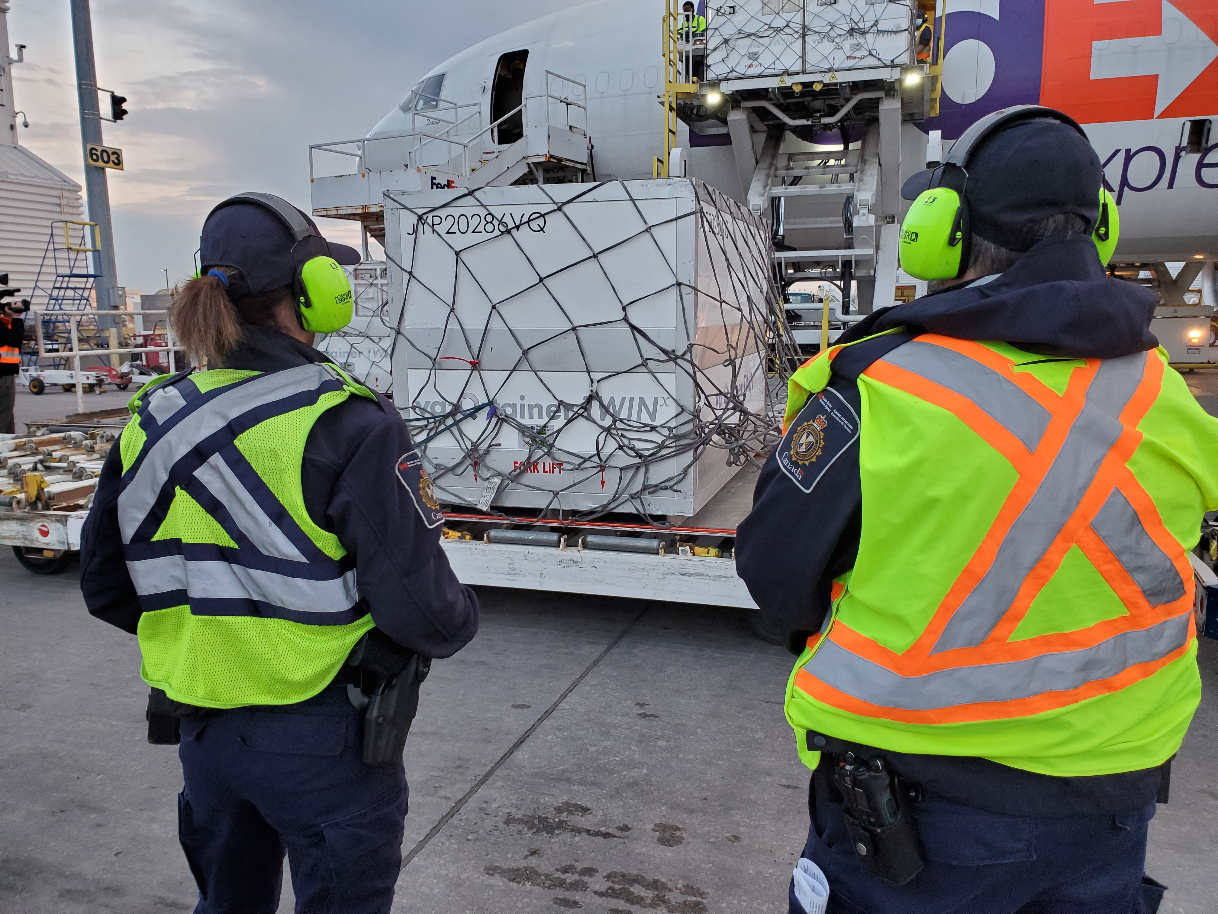 Employees in safety vests look on as a shipment of vaccines is lowered from a plane to the tarmac