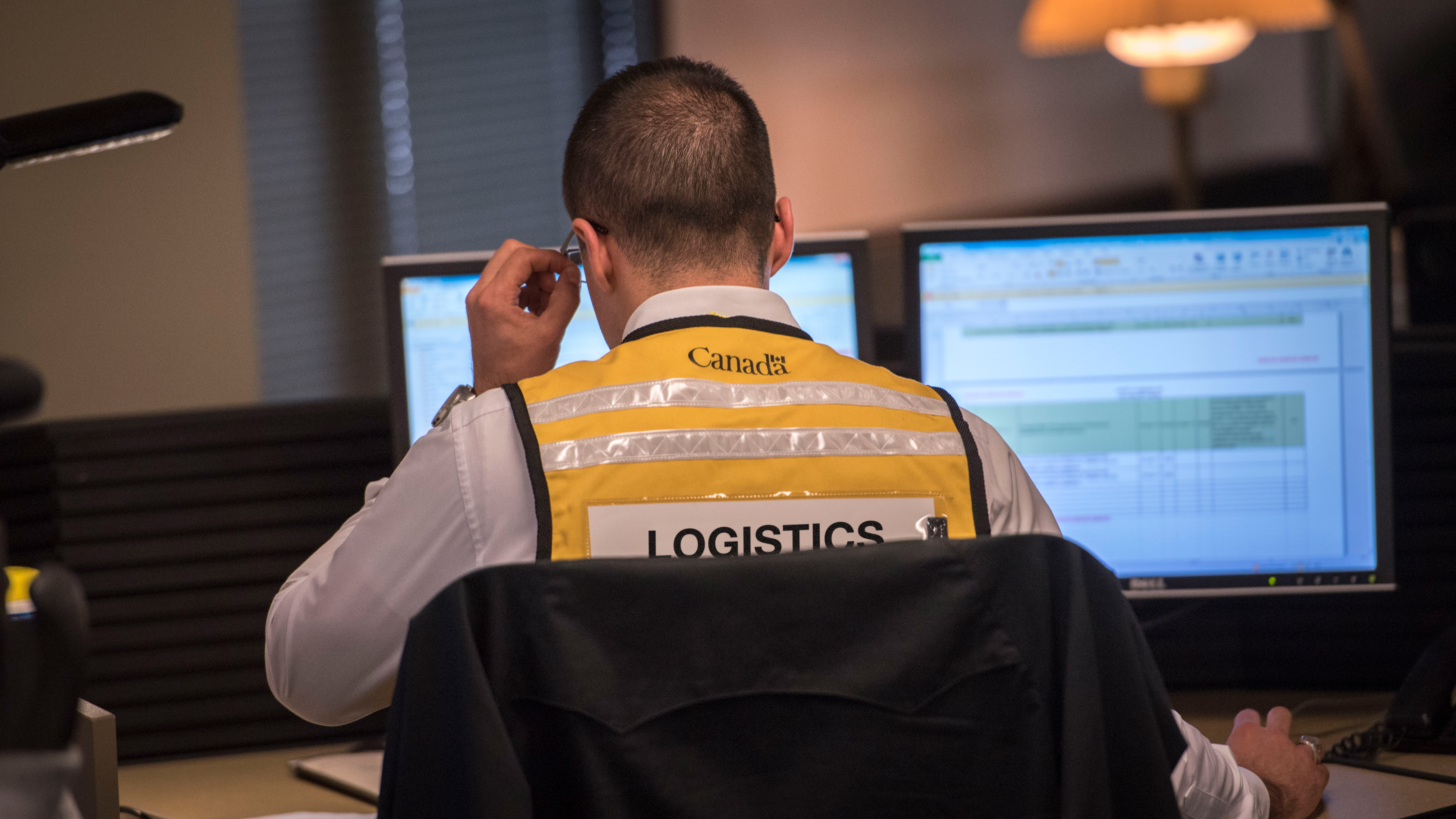 A public servant wearing a yellow vest that says “Logistics” on the back sits in front of a computer in the Government Operations Centre