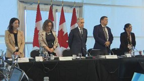 Mark Carney standing at a table with Ministers and senior government officials, two on either side of him. There are four Canadian Flags behind them