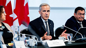 Mark Carney sitting at a table with a minister and senior government official on either side of him. There is a Canadian Flag behind them