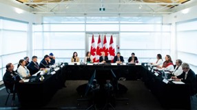 Government officials, ministers, and First Nations leaders sitting at tables in the shape of a horseshoe. Mark Carney sits in the center. There are four Canada flags behind the centre table