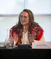 Mandy Gull-Masty speaking into a microphone using hand gestures while sitting at a table. There is a name card and a pitcher of water in font of her