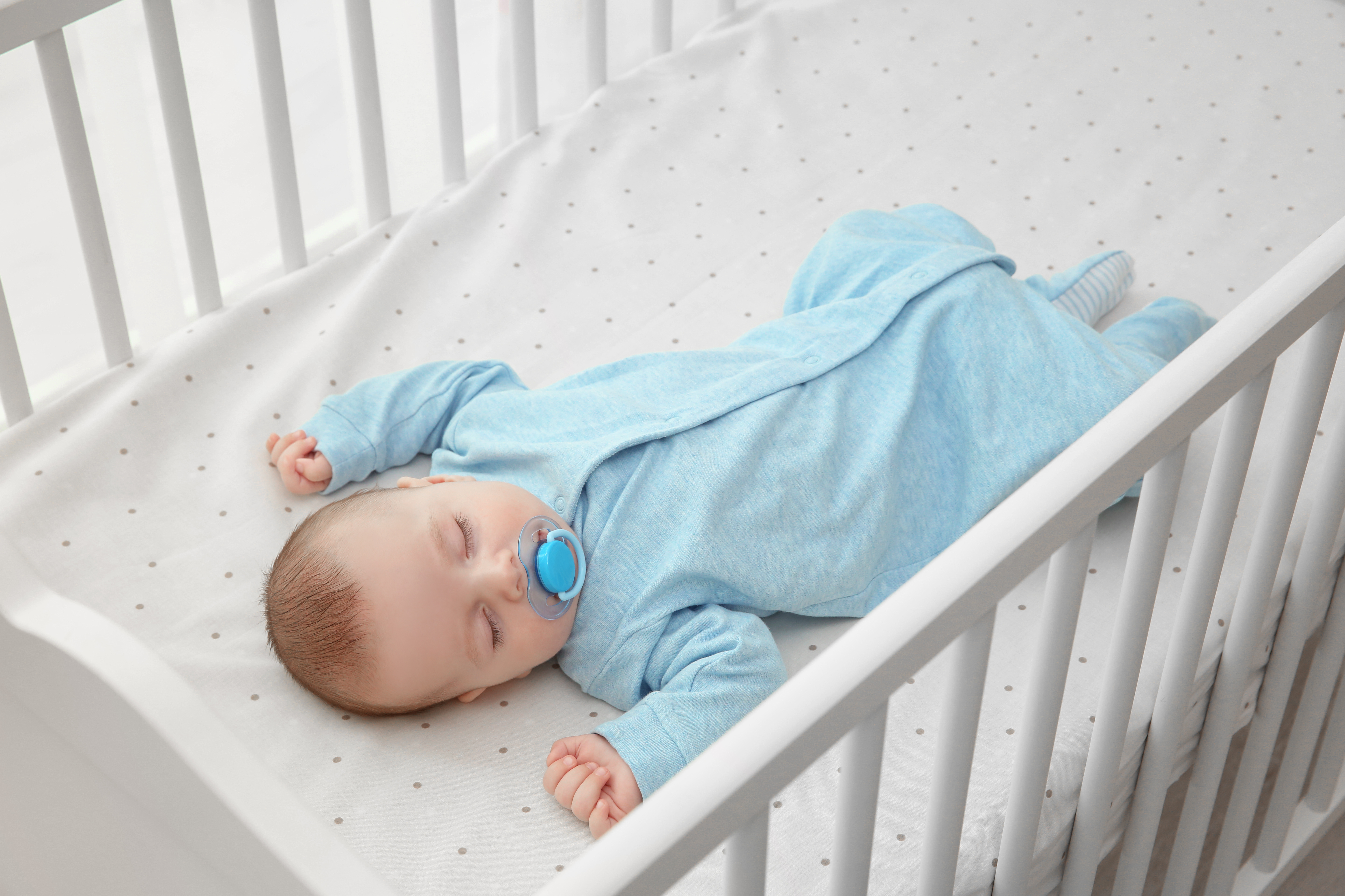 A baby asleep alone on his back in a bare crib. The baby is wearing one-piece sleepwear.
