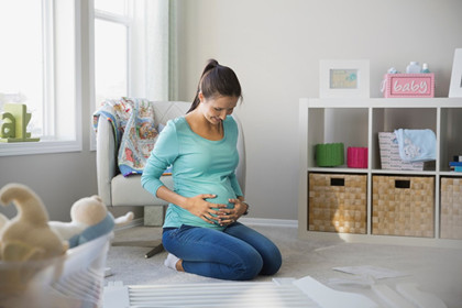 A pregnant person kneels in their child’s nursery with their hands on their belly