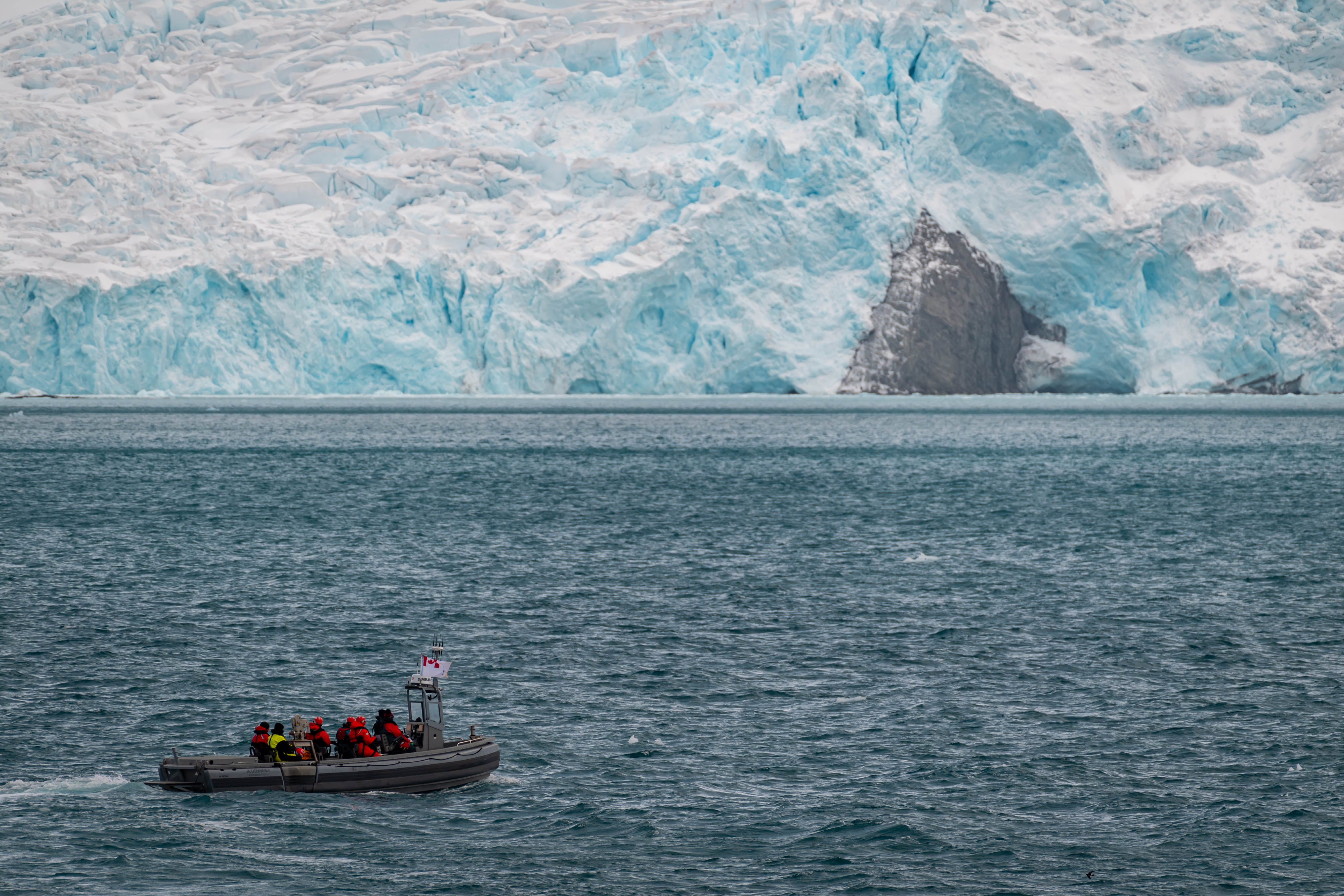 A large open boat with people in winter coats, cruising parallel to thick glacier ice.