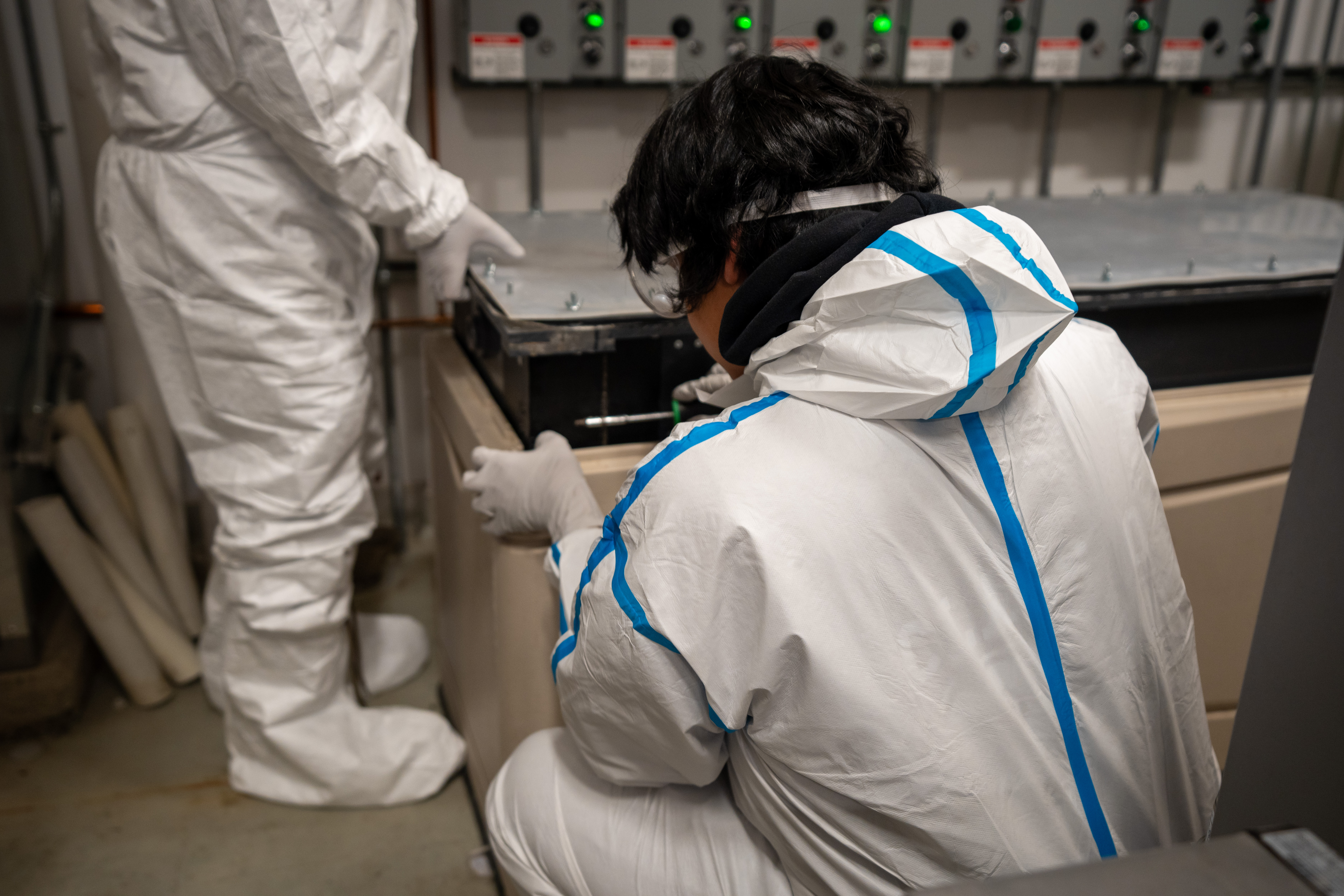The back of a man in a hazmat suit, crouching in front of a large box. To his left are a pair of legs in a hazmat suit.