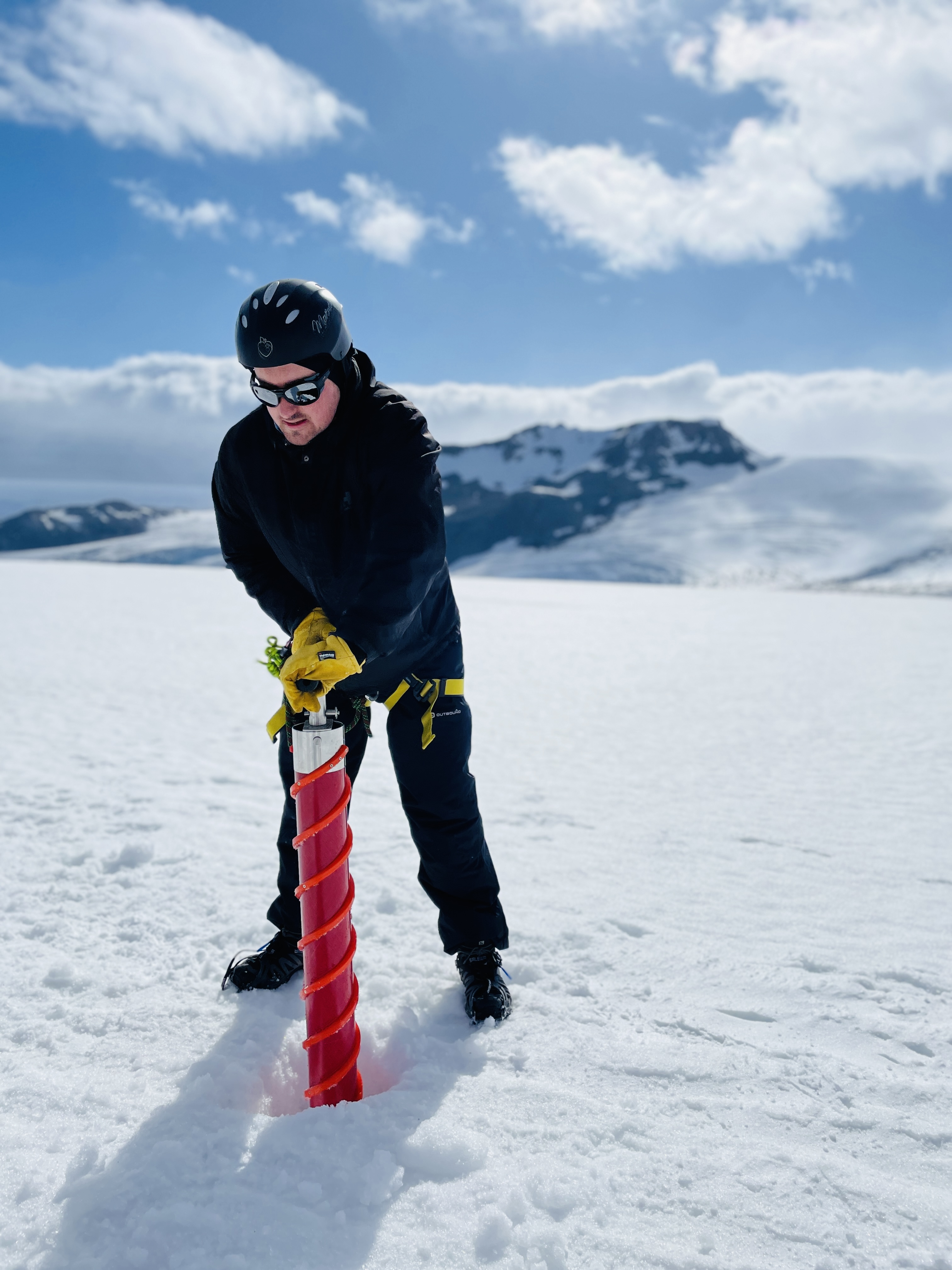 Man wearing a helmet, sunglasses, and winter clothing stands over a wide tube that he pulls out of the ice.