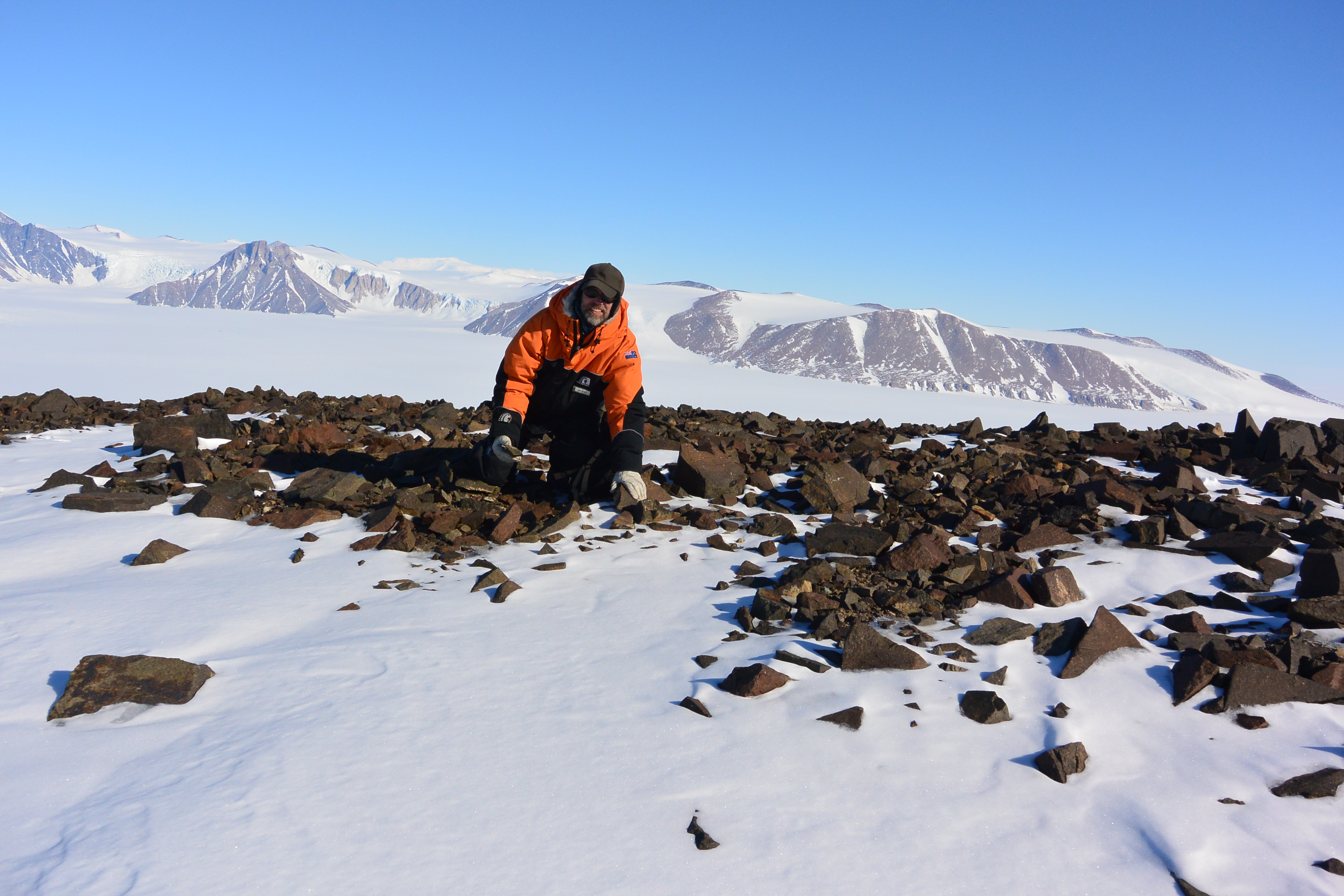 Man, wearing heavy winter jacket and hat, crouches on rocks. There are snow covered mountains behind him.
