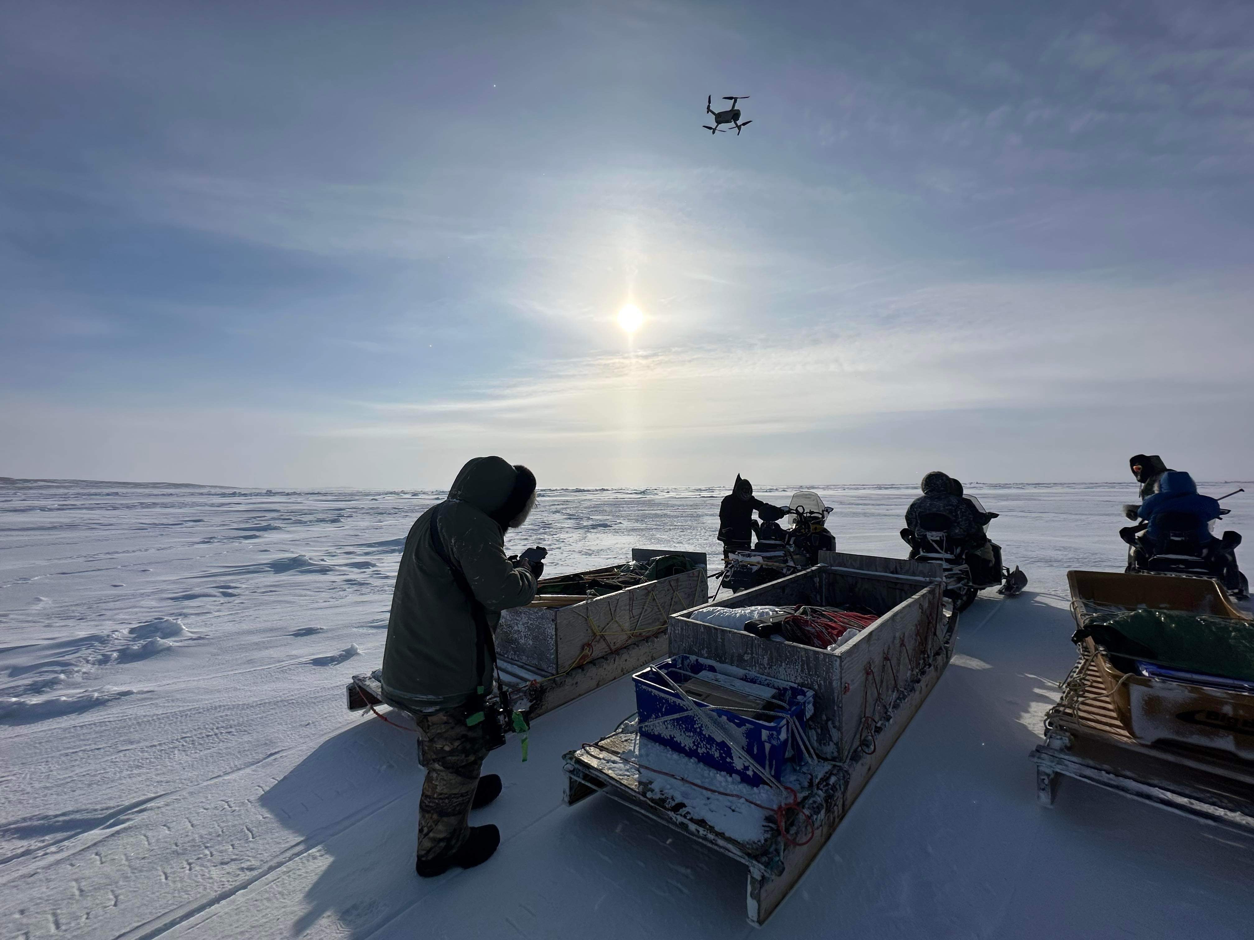 Three qamutiks and snowmobiles on snow facing away. Man in foreground operates a drone that is flying above their heads.
