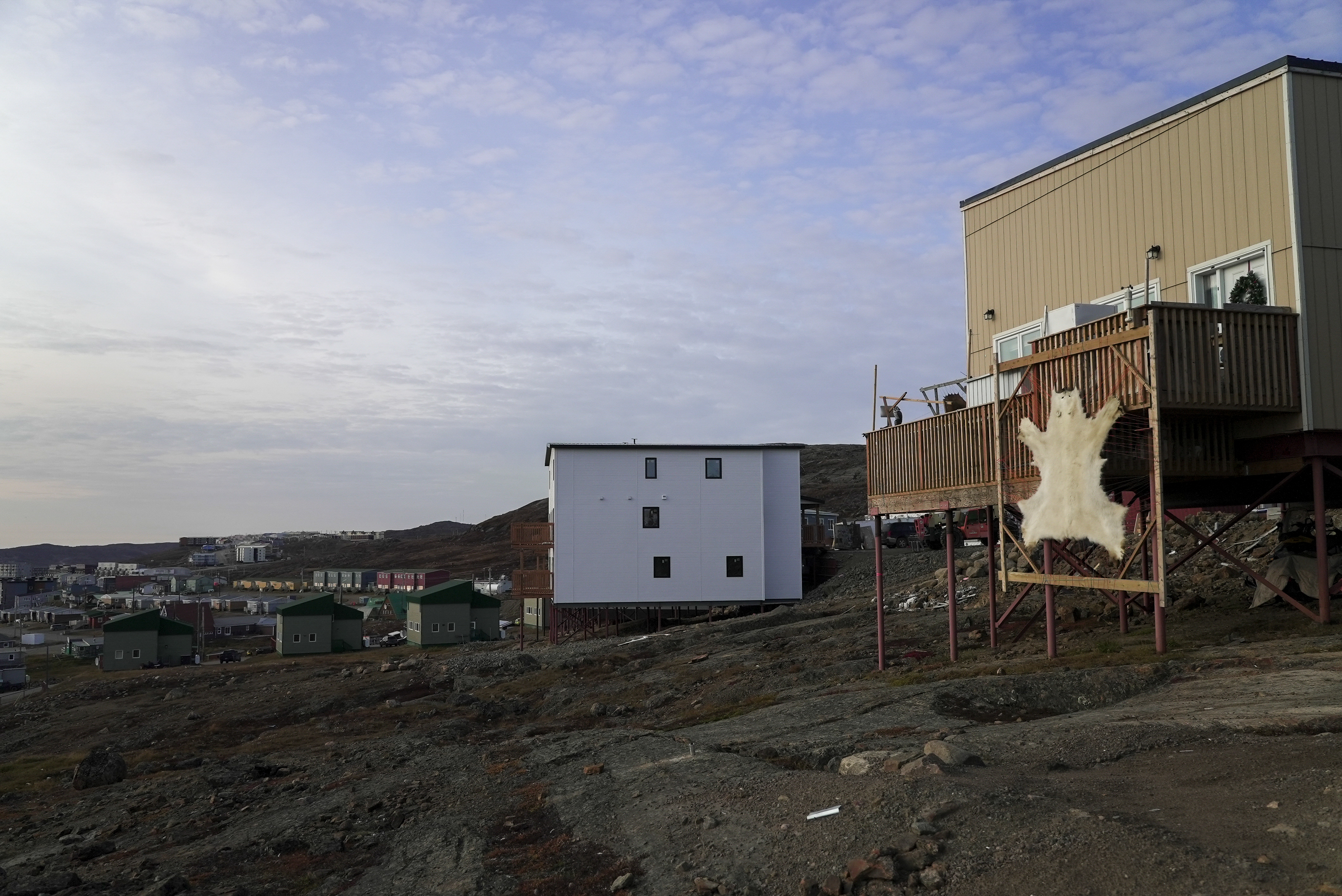 Two buildings, the one in the foreground has a high deck. A polar bear skin is stretched on a frame leaning against the deck railings.