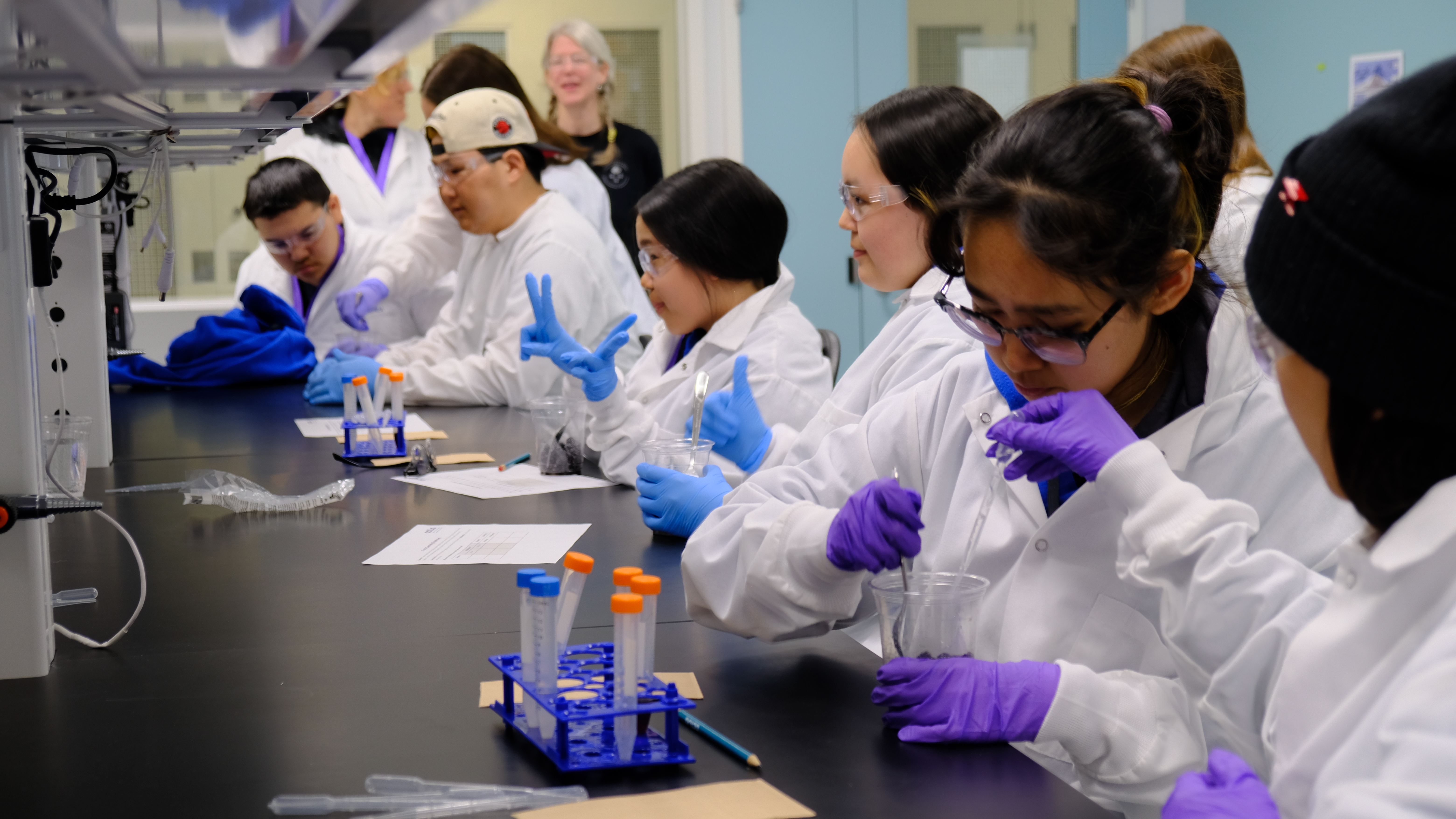 A row of teenagers in lab coats sit at a lab bench, work with test tubes.
